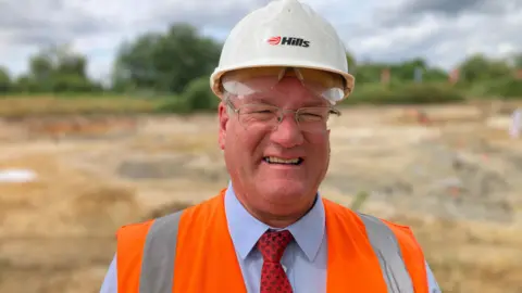 A man wearing a short and tie, an orange high vis vest and a white hard hat