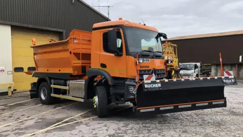 An orange gritter lorry parked outside of a warehouse on a cloudy day. There are two other vehicles parked nearby.