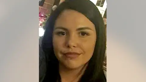 Alice Webb is smiling at the camera, she has dark hair and a piercing in her lower lip. She stands in front of an illuminated bar.