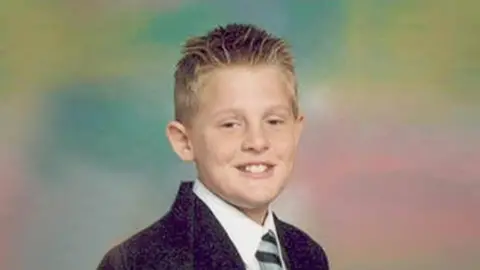 Lister family A child is dressed formally in a dark suit jacket, white shirt, and a striped tie. The background is a soft, multicoloured gradient with shades of green, yellow, and pink.
