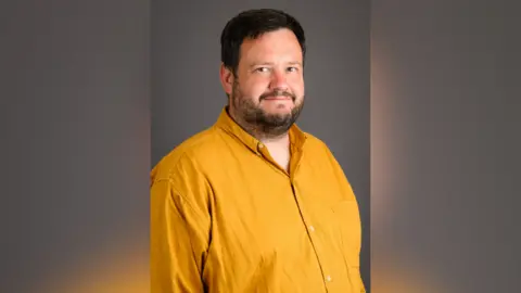 Bournemouth, Christchurch and Poole (BCP) Council A professional picture of a man wearing a yellow shirt. He is smiling into the camera with his body slightly tilted to the side.