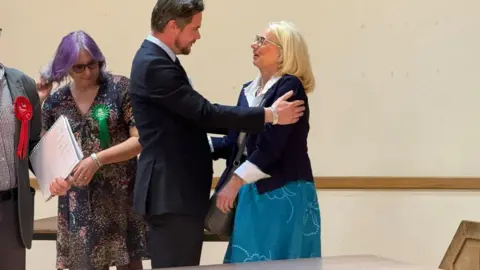 Louise Parker/BBC a man and a woman are standing next to each other and the man is holding the woman's shoulders smiling at her. They are at an election count and the woman has just won her seat, they are both wearing blue. 