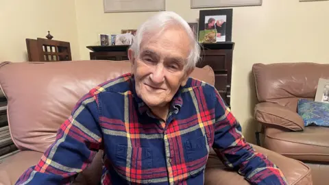 Suzy Lawson Noel Philo sits in a leather armchair looking towards the camera. He has white hair and is wearing a blue and red panelled shirt