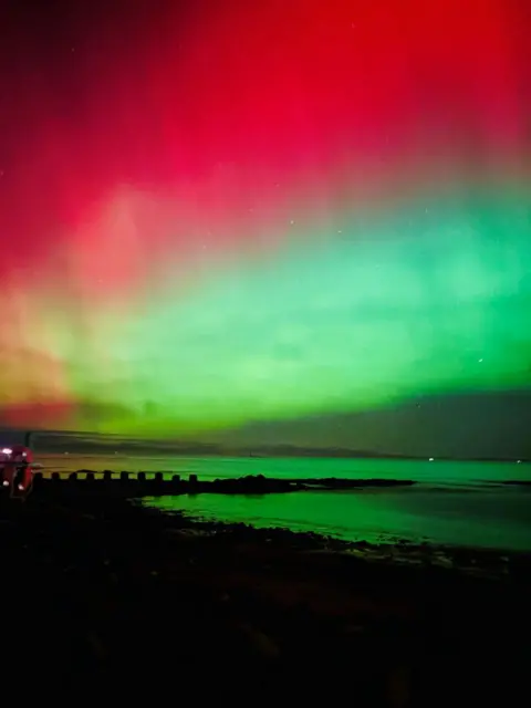 Ava Murchie Green and red aurora colours shine through cloud over a coastal landscape.