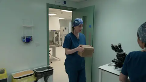 A woman in blue medical scrubs is seen carrying a plain box. She also is wearing a blue medical hair covering. She is in a room featuring a microscope, three bins typically found in a hospital, a light green door and turquoise walls. 