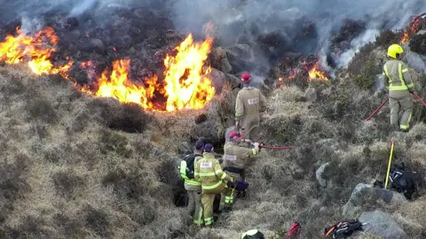 PA Media Five firefighters dressed in khaki uniforms battle flames on a gorse-covered slope of the mountains