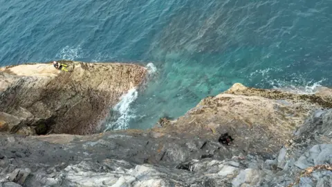 Boscastle Coastguard a photo from above showing two people sitting on rocks either side of a wide gully with sea between them