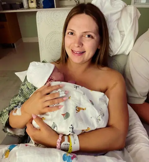 Maisie Savage Newborn baby wrapped in a white patterned hospital blanket and wearing a white knit hat, held against the chest of a person seated in a hospital bed. The person has a hospital wristband and an adhesive dressing on one wrist. White bedding and pillows surround the bed. A partially visible person in a white shirt sits beside them. In the background, a hospital room contains a sink, countertop and storage area with medical and personal items.