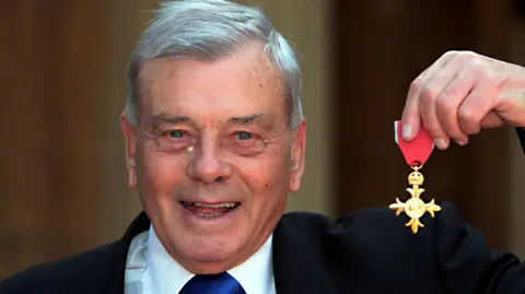 Reuters An older, grey haired gentleman poses happily with a medal of honour. He is Dickie Bird, the famed late cricket umpire, and he is posing with his OBE.