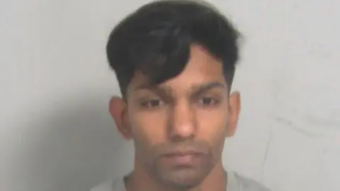 Essex Police Stephen Mahebadevan in a police custody picture. He has black hair and stubble and is wearing a grey sweater. He is expressionless.