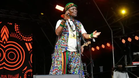 Redferns/Getty Images Emmanuel 'Jagari' Chanda sings into a microphone on stage, wearing a colourful outfit and hat
