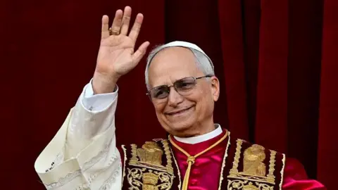 Getty A photograph of Pope Leo waving to crowds. He is wearing an embroidered gown with red, cream and gold accents. 