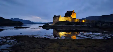 Viktor Tregubov A castle illuminated by artificial light taken at night. A loch and hills are visible in the photo.