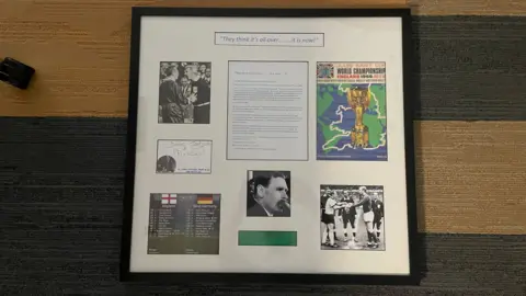 NICK WILSON/BBC This image shows a framed football (soccer) memorabilia display mounted against a textured wall with horizontal bands of beige and dark grey.
The frame is black, rectangular, and oriented horizontally.
Inside is a white background with a carefully arranged collage of images, text, and memorabilia related to a historic football event.
The layout is symmetrical, with items spaced neatly across the frame.