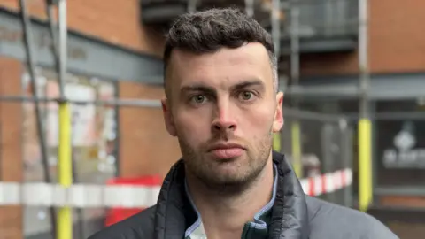 A man with brown hair and brown facial hair is standing in front of a building with metal scaffolding in front of it. He wears a dark top with a black puffer jacket on top