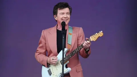 Rick Astley wears a pink suit and plays a guitar and stands at a microphone