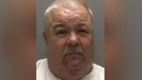 Police mugshot image of Gary Ward. He has a grey moustache and hair and is wearing a white top. He is pictured in front of a grey background and is squinting at the camera.