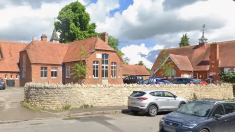 Google Two red brick buildings behind a Cotswold stone wall. There are two cars on the road and several parked in the car park in front of the buildings. It is a sunny day and there is a tree in the background.