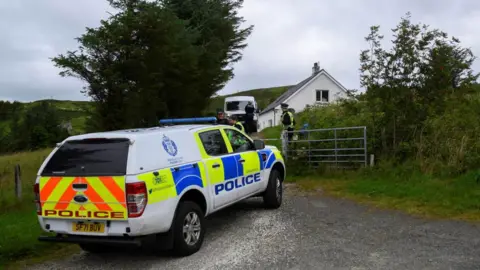 A police truck is parked at the entrance to the property. A police officer stands next to a metal farm gate and another vehicle and officer is parked near the house.