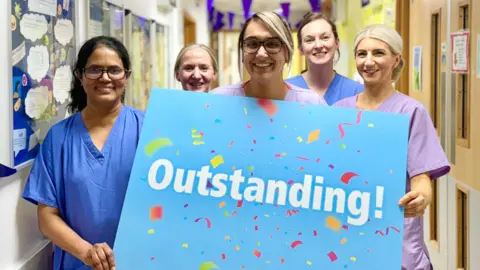 Five medical staff are dressed in scrubs and holding a blue banner saying the word outstanding! in a hospital corridor 