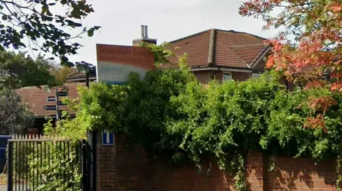 Google Stonedale Lodge is a red brick building set behind bushes and trees. A sign with black writing against an orange background reads 'Stonedale Lodge'