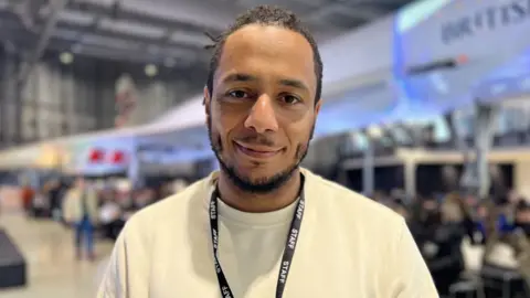 A midshot of Junior, smiling to camera, wearing a lanyard and a white jumper with the large hangar housing Concorde behind him