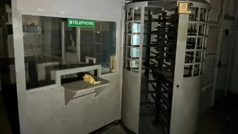 Shaun Whitmore/BBC A metal grey-painted turnstile gate and security office. A telephone is mounted on a shelf, beneath a window to the security office. A green sticker stating: "telephone" is stuck onto the glass.