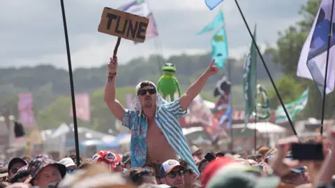 PA Media A man in an open blue and white shirt sits on the shoulders of a friend at Glastonbury festival with a sign that says "Tune". Kermit the Frog is held aloft behind him