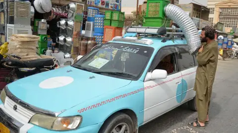 Getty Images Taxi driver Abdul Bari installing a hand-made cooler on top of his blue vehicle, in front of a store selling these customised air-cooling units