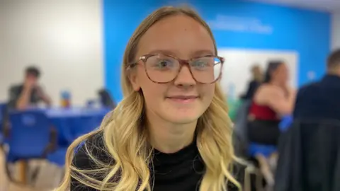 BBC Katie has long blond hair and wears large glasses. She smiles at the camera in the audition room.