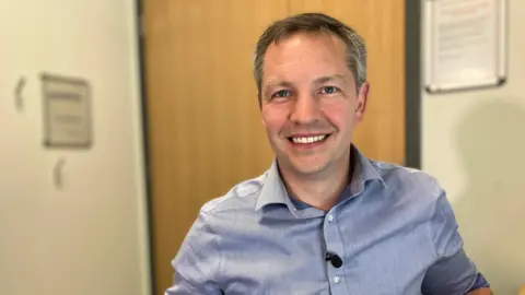 Richard Bamford wearing a blue shirt and smiling. He is in a doctor's office with a door behind him.