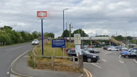 Google The image shows the entrance area to Thirsk railway station. A road curves gently uphill on the left, with a car travelling along it, and a tall British Rail-sign displaying the station name “Thirsk” on a pole beside the road. To the right is a large car park filled with vehicles, with marked bays and signs. A pedestrian footbridge spans the railway in the background, with stairs leading up to it. The surroundings include trees, grass, and a partly cloudy sky.
