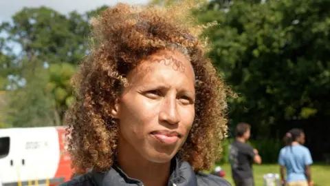 Serena looks at the camera as people set up the sports day behind her. She is wearing a white jumper with blue stripe hoops with a black gillet. Her hair is slightly blowing in the wind.