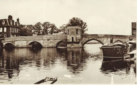 Norris Museum, St Ives St Ives town bridge in black and white from 1930, five arches can be seen crossing the river with a chapel in the middle with just one single building with arched windows in limestone.