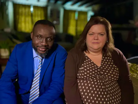 MARTIN GILES/BBC Caroline with her husband Lionel, both look at the camera. Lionel is on the left, wearing a royal blue suit with a blue, yellow and black striped tie. Caroline is wearing a brown spotted dress with a chocolate brown cardigan.