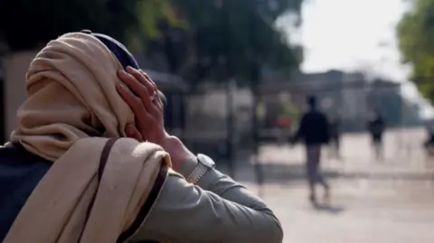 A woman who wears a brown head scarf heads away from the camera to some large doors. She is holding her head in her hands. 