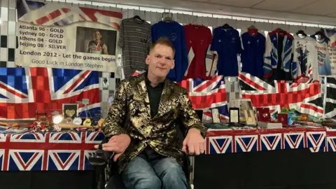 Stephen Miller, who is wearing a gold and black blazer, posing in front of his medals and memorabilia at a celebration of his career at St James' Park in Newcastle.