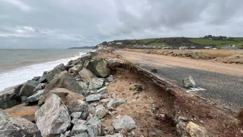 Road at Torcross with large hole undermining it. The sea is to the left. The sky is grey and cloudy. 
