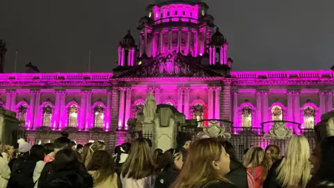 Belfast City Hall is illuminated by pink lights. It is night and hundreds of protesters are pictured in front of the building behind the black iron gates