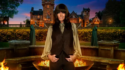 BBC/Studio Lambert/Cody Burridge/Matt Burlem Claudia Winkleman poses outside the Traitors castle in a promotional image for series four. There are two figures in green cloaks behind her also.