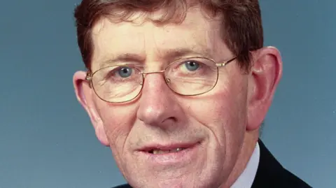 Thomas Gallagher has blue eyes, brown short hair and is wearing gold-rimmed glasses.
He is wearing a black suit and white shirt. 