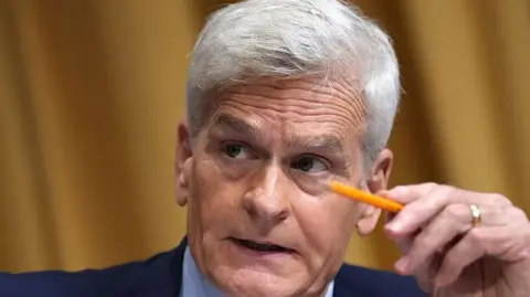 close-up of a white-haired Bill Cassidy sitting behind a desk and holding a pencil in his hand while talking