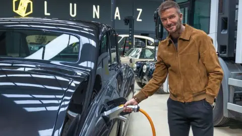 David Beckham wearing mustard-coloured jacket plugging charging lead into car