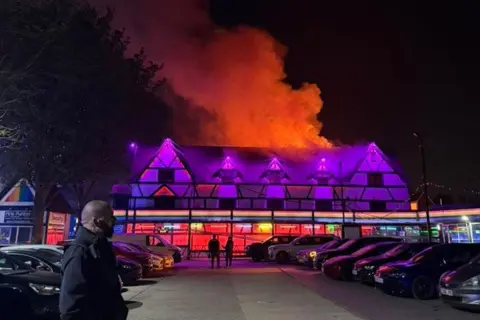 Gabby Cantareli The outside of Pink Punters, a night club in Fenny Stratford. There are purple neon lights and smoke and orange flames coming from the top of the building.