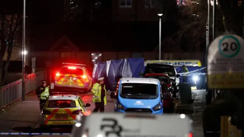 Stephen Gibson/Buchanan Photos A blue police tent has been set up in the middle of a road. It is dark and the street lights are on. Two police cars are parked on the side of the road and one of them has its lights on. Police officers walk down the road and the road is taped off.