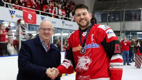 Swindon Wildcats A man with a short beard and wavy brown hair wearing a red hockey top shakes the hand of a man in a blue jumper as they stand together on an ice hockey rink. Spectators and other players are visible in the background