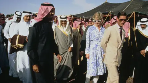 Tim Graham Photo Library via Getty Images Charles, wearing a cream linen suit and sunglasses, and Princess Diana, wearing a blue and white floral dress and sunglasses, walk along smiling, surrounded by many men in traditional Saudi attire, also smiling.