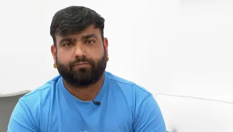 A antheral   with abbreviated  hairsbreadth  and a beard and moustache, wearing a bluish  t shirt. He is looking astatine  the camera but is not smiling.