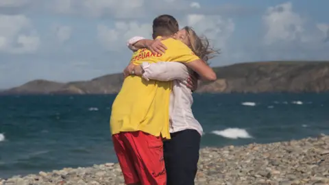 Matty and Joanna hugging on Newgale beach. Matty's face isn't visible as he's hugging Joanna. He is wearing his RNLI uniform - a yellow t-shirt and red shorts. Joanna's face can be seen, her eyes are shut and her long blonde hair is blowing in the wind. She is wearing a light pink long sleeve shirt and black trousers. 