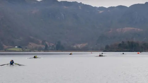 A number of crews in rowing boats are rowing across Derwentwater - a very large lake in the Lake District. The lake is surrounded by mountains. 